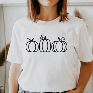 以下が含まれることがあります： 白いTシャツに、黒い輪郭で描かれた3つのカボチャのイラスト