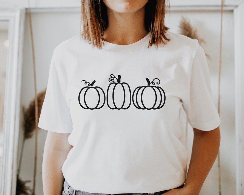 以下が含まれることがあります： 白いTシャツに、黒い輪郭で描かれた3つのカボチャのイラスト
