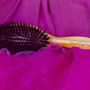 以下が含まれることがあります： 黒い毛のついた木製のヘアブラシのクローズアップ。ブラシはピンクのキラキラした布の上に置かれています。ハンドルには「Head Wrap Nest」という文字が刻まれています。