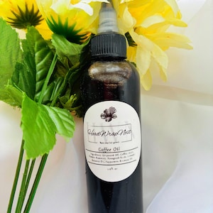 May include: A dark brown bottle of Head Wrap Nest Coffee Oil with a black cap and white label. The label includes the product name and ingredients. The bottle is surrounded by yellow flowers and green leaves.
