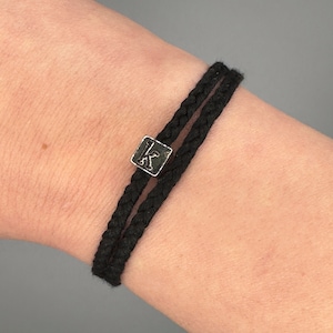 May include: Black braided bracelet with a silver square charm bearing the letter "K". The bracelet is made of two parallel strands of black cord. The charm is positioned centrally.