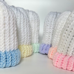 May include: A collection of white knit baby hats with colorful pastel brims. The hats have a ribbed texture and are displayed in a row, showcasing the blue, yellow, green, purple, and pink accents at the base.