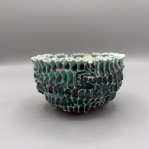 May include: A small ceramic bowl featuring a highly textured, honeycomb-like exterior surface. The glaze is a blend of deep teal and dark charcoal tones, creating a rugged, organic appearance with a smooth, light-colored interior rim.