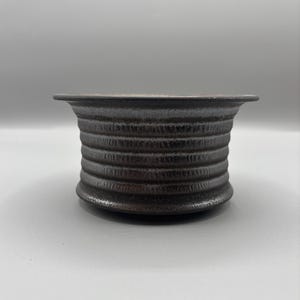 May include: A dark brown, textured ceramic bowl with a wide, flat rim. The bowl's body features horizontal, ridged detailing, creating a ribbed effect. The interior is a darker shade, contrasting with the exterior. The bowl sits on a light gray surface.