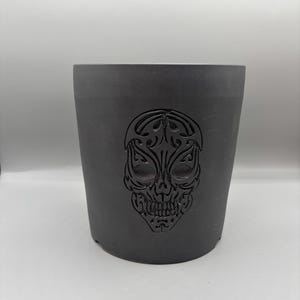 Może przedstawiać: Czarna ceramiczna doniczka z wygrawerowanym na przodzie wzorem czaszki.