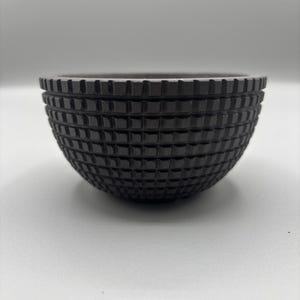 May include: A dark gray bowl with a textured, grid-like pattern. The bowl has a rounded shape and a slightly flared rim. The surface has a matte finish, and the bowl is set against a white background.