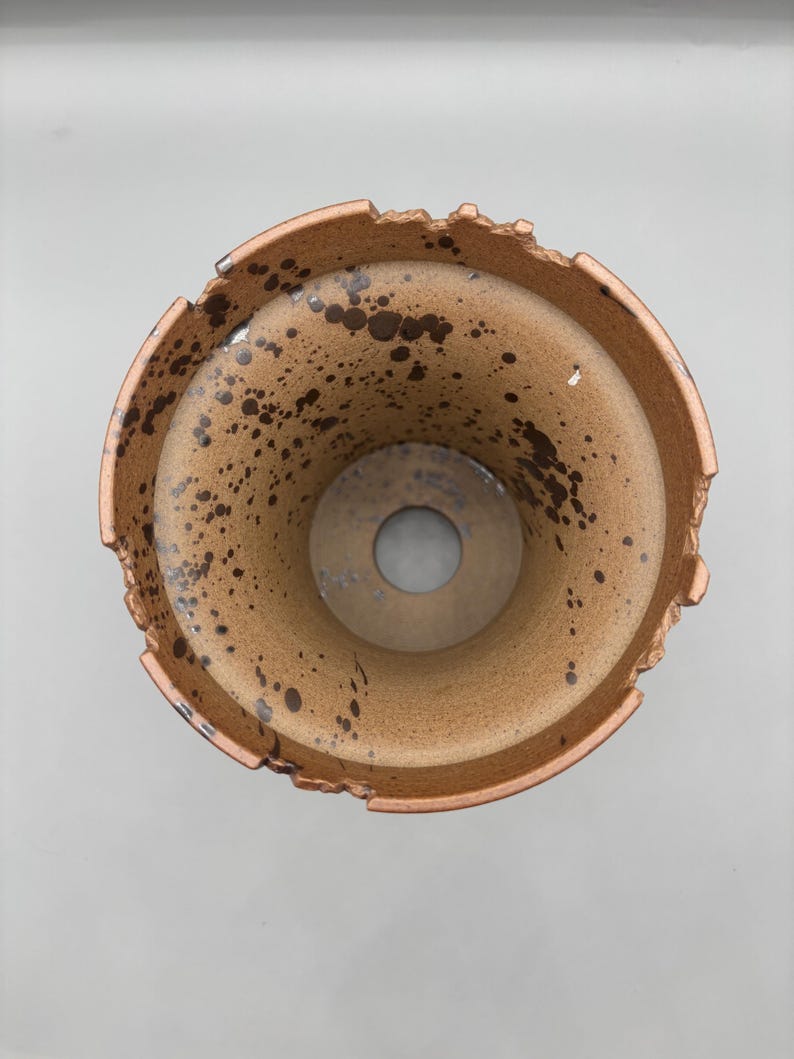 May include: A ceramic planter with a speckled, light brown interior and exterior. The rim has a rough, textured edge. Dark brown spots are scattered across the surface, adding a unique, rustic aesthetic. The planter has a circular hole at the bottom.