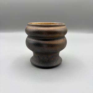 May include: A small, rounded ceramic cup with a dark, metallic bronze and black textured finish. The vessel features a distinct double-bulge silhouette with a flared base and a smooth, lighter-toned interior rim, showcasing a rustic aesthetic.