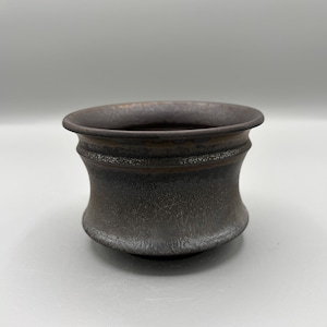 Peut inclure: Un bol en céramique brun foncé avec une surface texturée. Le bol a une forme large et arrondie avec un bord légèrement évasé. L'extérieur a un aspect moucheté et l'intérieur est d'une teinte plus foncée.