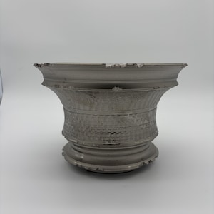 May include: A weathered, gray decorative planter with a wide, fluted top and a textured, cylindrical body. The planter has a distressed appearance, with chipped edges and a worn finish, suggesting an aged aesthetic. The base is a series of stacked rings.