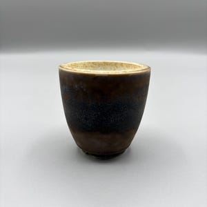May include: A small, handcrafted ceramic cup with a unique, textured surface. The cup features a gradient of colors, transitioning from a light beige rim to a dark brown base with a band of black in the middle. The cup has a simple, rounded shape.