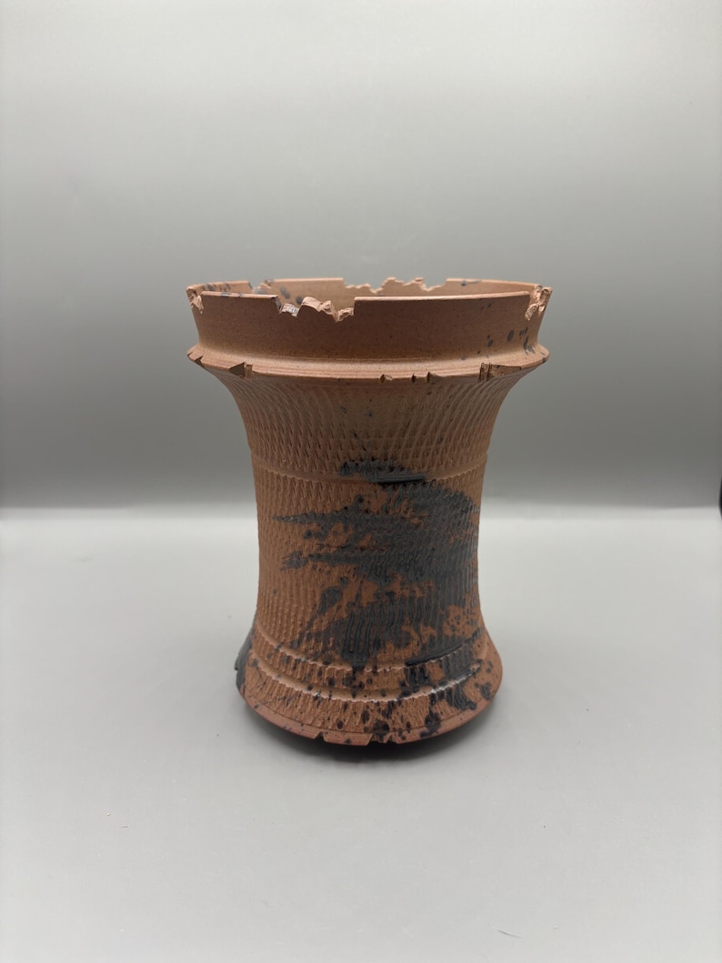 May include: A handmade, terracotta-colored ceramic vase with a textured, ribbed design. The vase features a unique, jagged rim and dark brown, abstract paint splatters. The vase is a decorative item.
