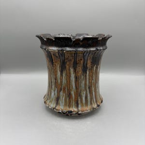 May include: A ceramic vessel with a flared, scalloped rim and a cylindrical body. The surface features a reactive glaze with vertical drips in shades of dark brown, metallic bronze, and muted blue, creating a textured, multi-layered appearance.