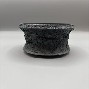 Puede incluir: Un cuenco de cerámica gris oscuro y texturizado con una base ancha y redondeada y un borde acampanado. El cuenco tiene una textura rugosa y orgánica, y el color es un carbón profundo. El cuenco es probablemente hecho a mano.
