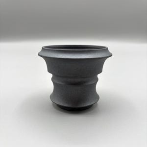 May include: A dark gray, decorative ceramic pot with a unique, layered design. The pot has a wide, flared top and a textured surface. The overall shape is reminiscent of a classic urn, suitable for displaying plants or as a decorative accent.