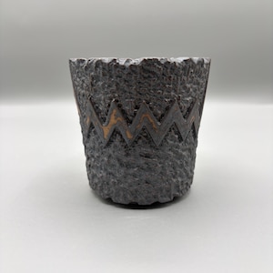 May include: A dark brown, cylindrical cup with a rough, textured exterior surface. The cup features a prominent, horizontal zigzag band around the middle, revealing a lighter, metallic bronze-colored finish within the recessed pattern.