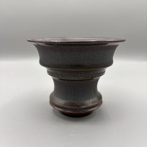 May include: A handcrafted ceramic vessel with a dark, mottled glaze. The piece features a wide, flared rim and a bulbous, pedestal-like base. The color palette includes shades of brown, black, and hints of red, creating a rustic aesthetic.