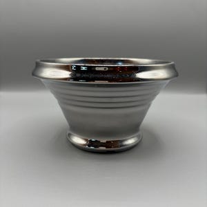 May include: A metallic silver bowl with a polished, reflective finish. The bowl features a wide, flared rim and a series of horizontal ridges encircling the body, tapering down to a circular pedestal base with a smooth, rounded edge.