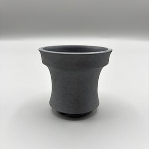 May include: A small, dark gray ceramic vessel with a textured surface. The cup has a flared rim and a rounded base. The design is simple and modern, suitable for a variety of uses.