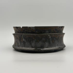 May include: A circular ceramic ashtray with a dark, mottled finish featuring shades of charcoal, black, and metallic bronze. The exterior has a concave, hourglass-like profile with a textured, uneven rim and a wide, flat base for stability.