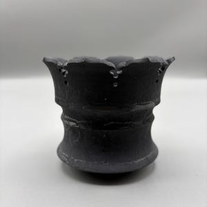 May include: A dark gray ceramic pot with a scalloped rim and a textured surface. The pot has a bulbous base and a cylindrical midsection. The rim has small, evenly spaced holes. The pot is set against a white background.