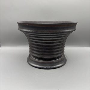 Puede incluir: Un cuenco de cerámica marrón oscuro, acanalado, con un borde ancho y una base acampanada y texturizada. El cuenco tiene una forma cilíndrica con crestas horizontales, creando un diseño decorativo único. El cuenco es adecuado para servir o exhibir.