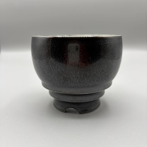 May include: A small, dark gray ceramic bowl with a speckled, metallic finish. The bowl features a rounded upper body that tapers down to a stepped, tiered base with a small cutout notch at the bottom edge, creating a distinct, layered profile.