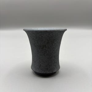 May include: A dark gray ceramic cup with a flared top and a rounded base. The cup has a textured surface and a smooth rim. The cup is designed with a minimalist aesthetic and is suitable for beverages.