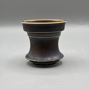 May include: A small, dark ceramic cup with a flared rim and a tapered, hourglass-shaped body. The exterior features a deep, mottled black and dark brown glaze with a textured finish, while the rim and base edges are highlighted with a thin, light brown line.