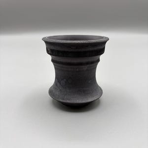 May include: A dark gray, hourglass-shaped decorative object. The object has a wide top and base, with a narrower, textured midsection. The surface appears to have a slightly rough texture. The object is likely made of ceramic or a similar material.