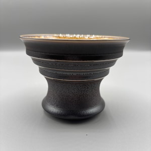 May include: A dark brown ceramic bowl with a textured surface and a gold-colored rim. The bowl has a unique, tiered design with a wide opening and a rounded base. The interior appears to be a lighter shade of brown.