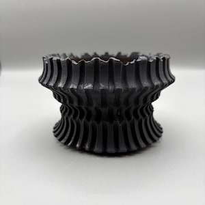 May include: A black, decorative bowl with a unique, textured design. The bowl has a fluted, wavy rim and a layered, ridged body. The dark color and intricate pattern create a striking visual effect, suitable for home decor.