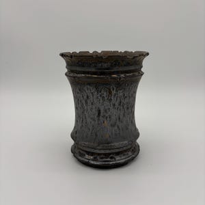 Peut inclure: Un vase en céramique avec un bord évasé et un corps fuselé, présentant une glaçure aux tons métalliques sombres avec une finition texturée et tachetée. La surface affiche un mélange de teintes charbon, bronze et cuivre avec un aspect brillant.