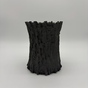 May include: A matte black cylindrical vase featuring a deeply textured exterior surface that mimics the rugged, vertical ridges and grooves of tree bark. The top edge is uneven and jagged, following the organic pattern of the textured sides.
