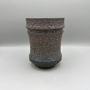 May include: A cylindrical ceramic cup featuring a heavily textured, porous surface with a dark charcoal grey and bronze-toned finish. The vessel has a slightly flared rim and a tapered base, showcasing a rugged, granular appearance throughout the exterior.