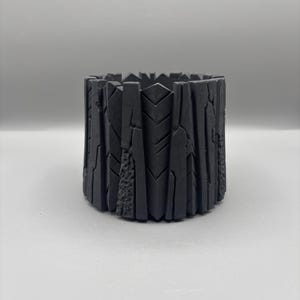 May include: A dark gray, cylindrical planter with a textured, bark-like exterior. The planter has a geometric, angular top edge and a rough, uneven surface, suggesting a natural, handcrafted aesthetic. Ideal for small plants or succulents.