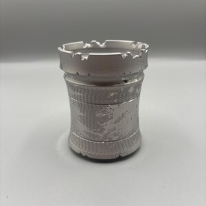 May include: A silver-colored ashtray with a castle turret design. The ashtray has a cylindrical body with decorative etched bands and a crenelated rim. The metal has a slightly distressed appearance.