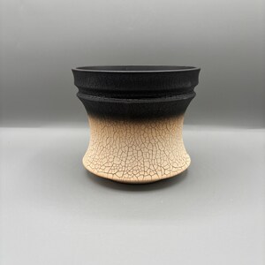 May include: A handcrafted ceramic vase with a unique design. The top portion is black, transitioning to a beige, cracked texture at the base. The vase has a flared shape and a textured surface, suitable for home decor.