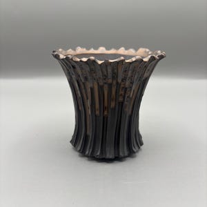 Puede incluir: Un jarrón decorativo de color marrón oscuro con un exterior acanalado y texturizado. El borde tiene un borde festoneado con un acabado metálico más claro. El jarrón tiene una forma ancha y acampanada.