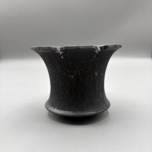 May include: A dark gray, textured, flared vase with a wide, uneven rim. The vase has a rounded base and a cylindrical body that widens towards the top. The surface appears to have a rough, almost pitted texture. The background is a plain, light gray.