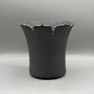 Puede incluir: Un recipiente cilíndrico de color negro mate con un borde superior acampanado que presenta un diseño festoneado. El borde está decorado con pequeños recortes espaciados uniformemente en la base de cada festón, creando un patrón detallado.