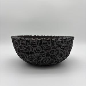 May include: A dark gray bowl with a textured, wave-like pattern. The bowl has a rounded shape with a scalloped rim. The surface of the bowl is covered in a repeating design of curved lines.