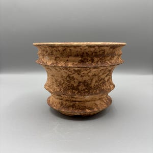May include: A brown ceramic planter with a textured surface. The planter has a wide, open top and a rounded base. The exterior features a speckled pattern and a series of horizontal ridges, giving it a rustic appearance.