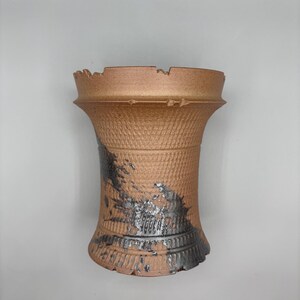 May include: A handmade, textured ceramic vase with a unique, distressed design. The vase features a warm, earthy brown color with accents of black and silver. The top edge has an uneven, jagged design.