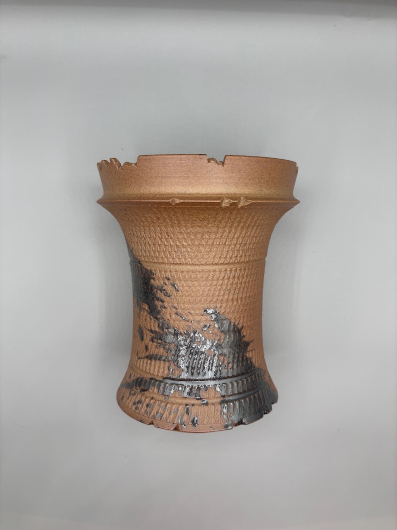 May include: A handmade, textured ceramic vase with a unique, distressed design. The vase features a warm, earthy brown color with accents of black and silver. The top edge has an uneven, jagged design.