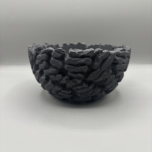 May include: A dark grey bowl with a textured, stone-like finish. The bowl has a rounded shape and an uneven rim, with a surface designed to resemble stacked stones. This decorative bowl is suitable for home decor.