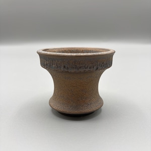 May include: A small, brown ceramic vessel with a textured surface. The vessel has a wide, slightly flared rim and a rounded base. The body of the vessel has a hourglass shape. The rim and the base have a darker brown color.