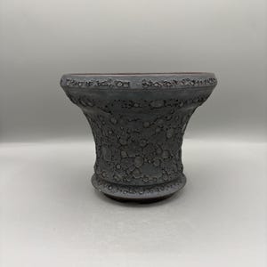 May include: A dark gray ceramic planter with a wide, flared rim and a tapered base. The exterior surface features a distinct, cratered, and textured finish that resembles a lunar landscape, with varying shades of charcoal and matte gray tones.