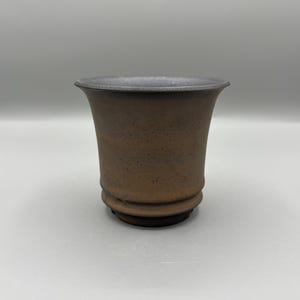 Peut inclure: Un pot en céramique marron avec un dessus évasé et une surface texturée. Le pot a un bord foncé et des rainures horizontales près de la base. La forme est un cône tronqué. Environ 10 cm de hauteur.