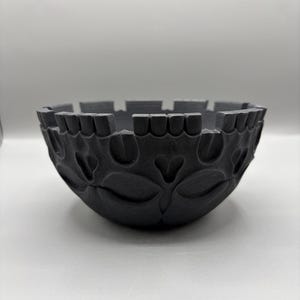 May include: A matte black bowl with a textured exterior featuring a repeating pattern of embossed heart-shaped motifs. The rim of the bowl is constructed with a series of evenly spaced, rectangular vertical segments that create a notched edge.